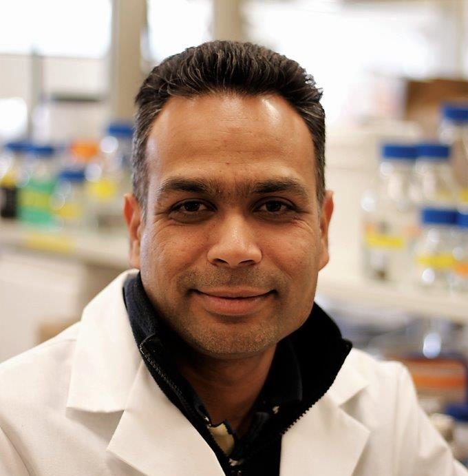 headshot of an indian man in a white coat, with a lab in the background
