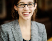 Une photo d'une femme blanche à lunettes et un blazer gris, souriant