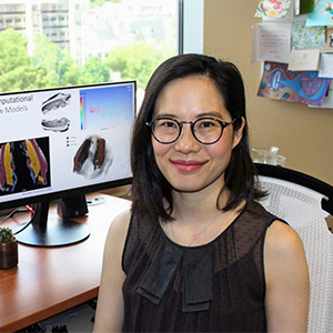 photo of Nicole Yee-Key Li-Jessen with glasses sitting in front of a computer monitor with slides showing her research