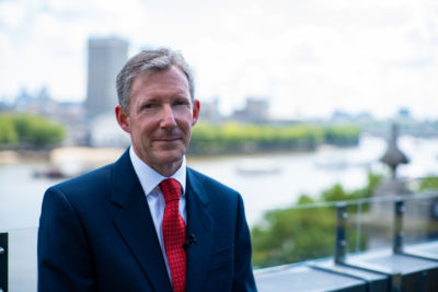 Photo of a white man in a suit on a balcony with the background blurred out.