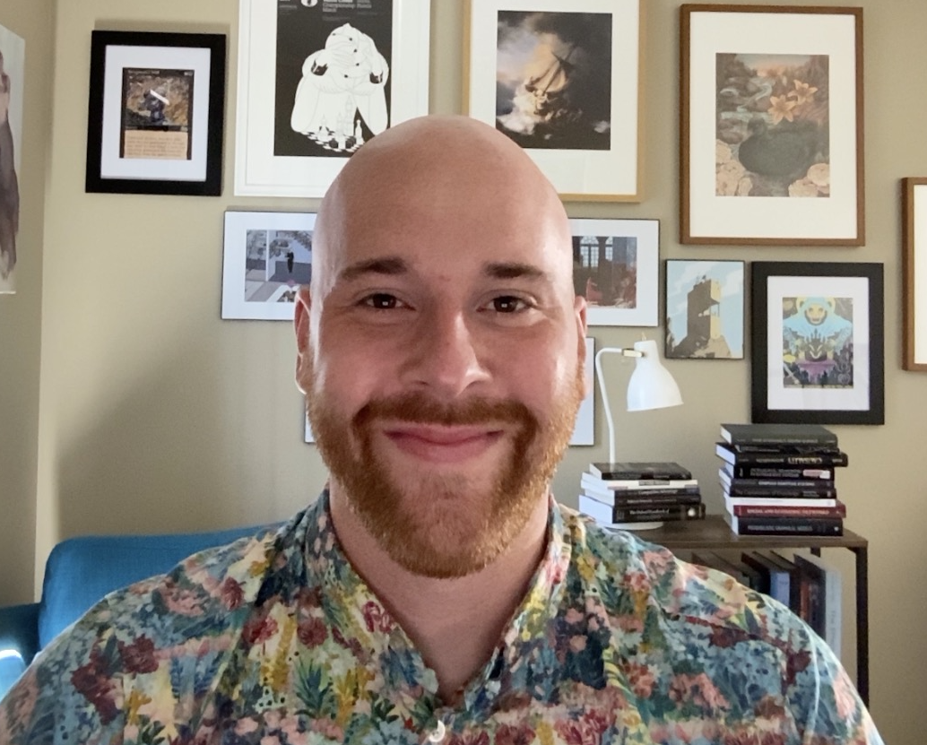 Headshot of a bald white man with a goatee. The wall behind him has many small pciture frames.