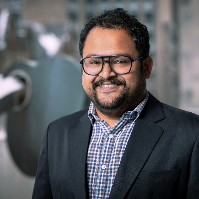 A headshot of an Indian man in a suit and glasses smiling at the camera.